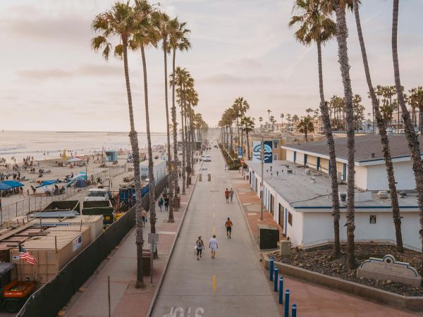 A palm-lined beachfront walkway with people, buildings, and umbrellas along the ocean under a hazy sky.