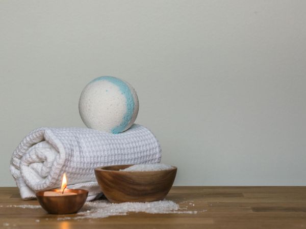 A bath-time scene with a blue bath ball, rolled towels, a lit candle, and a wooden bowl on a table.