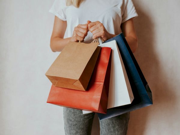 A person holding several shopping bags&mdash;brown, orange, blue, and white&mdash;filled with purchases while standing against a light background.
