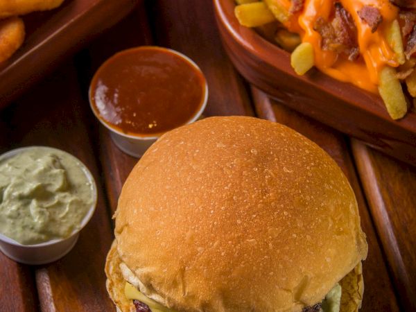 A burger sits on a wooden board with fries, onion rings, and two dipping sauces, all ready for a messy bite.