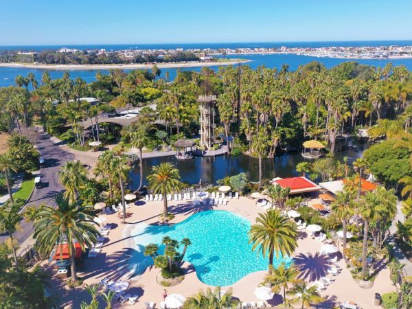 A sunny resort with a large turquoise pool, palm trees, shaded lounge areas, and a calm waterfront in the background.
