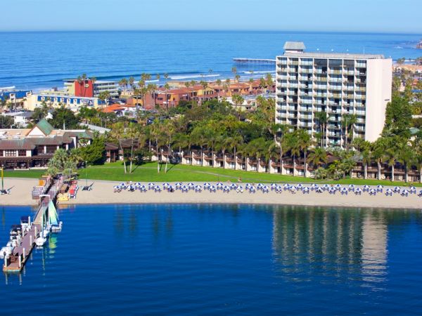 A coastal resort with a beachfront hotel, palm trees, lawns, rows of chairs, and a pier over calm blue water.