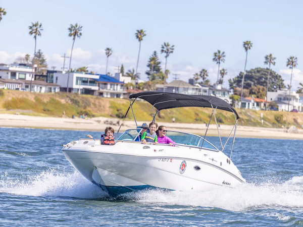 A white motorboat with a black canopy speeds across blue water, three people smiling aboard, palm trees and houses lining the shore in the background.