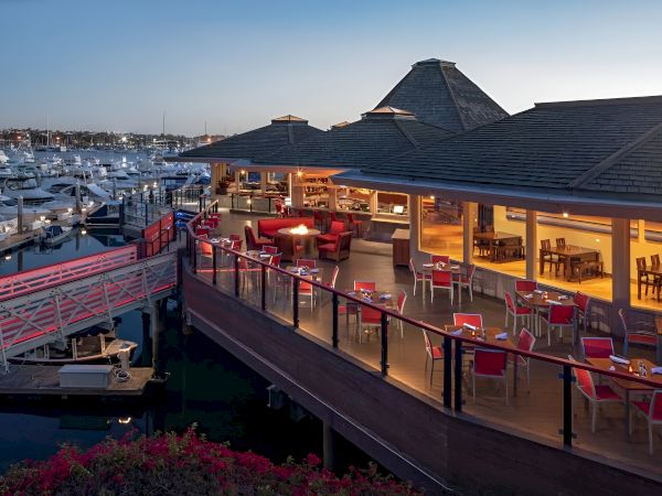 A seaside restaurant on a pier with outdoor seating, red chairs, warm lighting, and boats docked nearby, sunset sky.