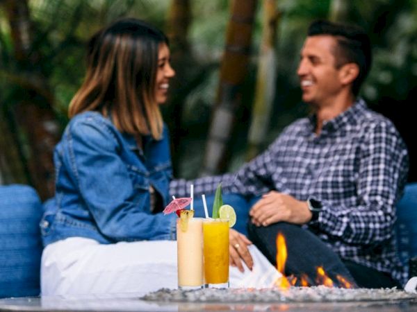 Two people chat and laugh on a blue outdoor couch by a fire pit, with colorful drinks in the foreground. End the sentence.