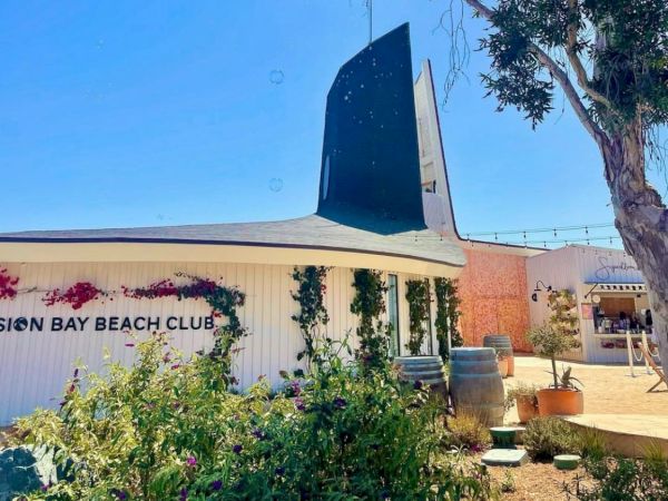 A mid-century clubhouse with a curved roof, sign &ldquo;Sun Bay Beach Club,&rdquo; modernist vertical sail sculpture, sunny courtyard, and landscaping.