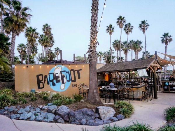 An outdoor bar and dining area named Barefoot Bar & Grill, with palm trees, string lights, and a sandy, relaxed tropical vibe.