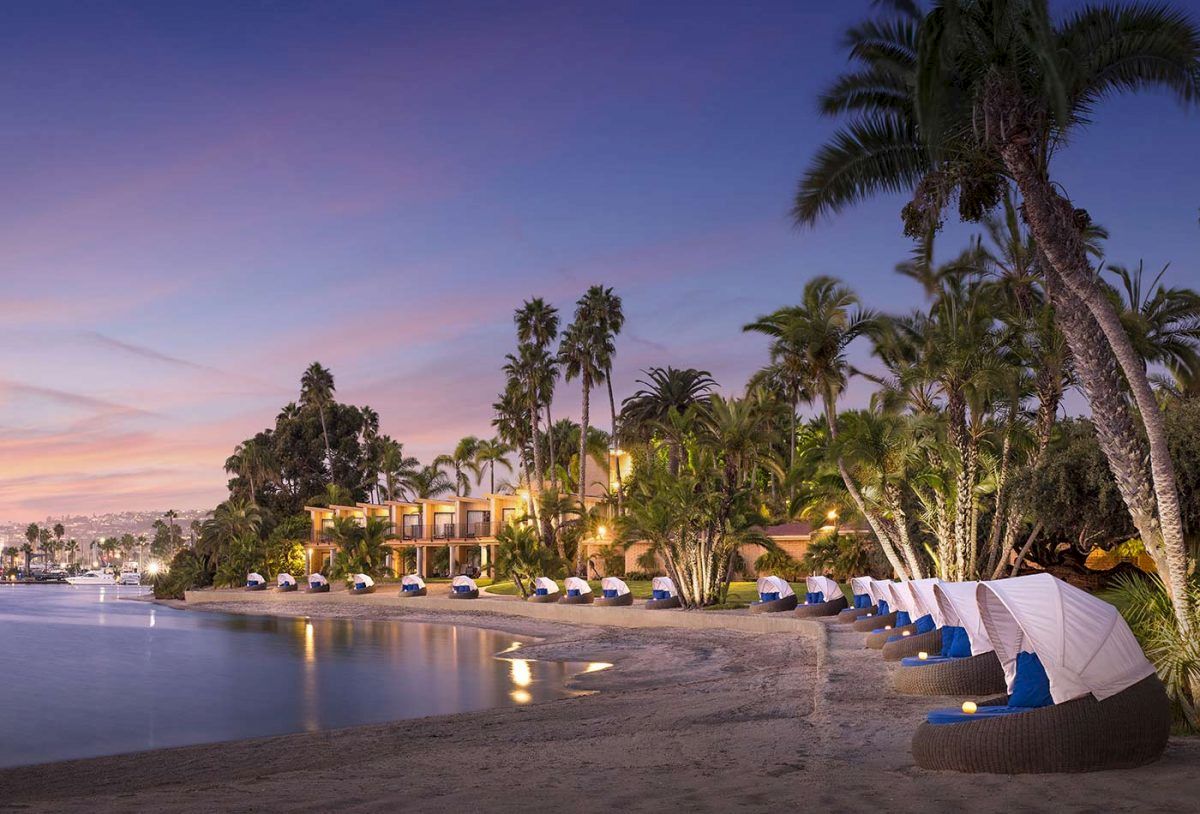 A tropical resort beach at twilight, palm trees, calm water, luxury huts lined along the shore with soft lighting.