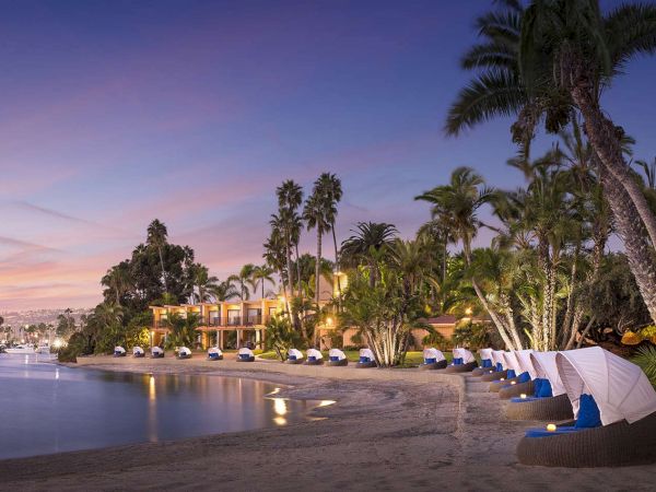 A tropical beachfront resort at dusk with palm trees, shaded cabanas, lounge chairs along the sandy shore, and calm water reflecting the sunset light.