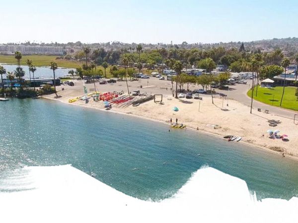 A sandy beach with calm blue water, boats on the left, palm trees, people sunbathing, and a park area with green grass alongshore, under a clear sky.
