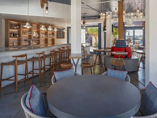 A modern lounge and bar area with round tables, wicker chairs, a long wooden bar, warm lighting, and a casual, airy atmosphere.