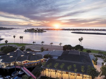 Sunset over a marina with palm trees, water, and a large seaside building complex, glowing lights reflecting on the calm harbor.