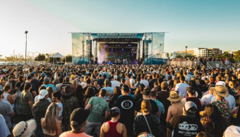 A large outdoor concert crowd facing a stage, sunlit, many hats and casual wear, festival vibe with a clear sky and skyline in the distance.