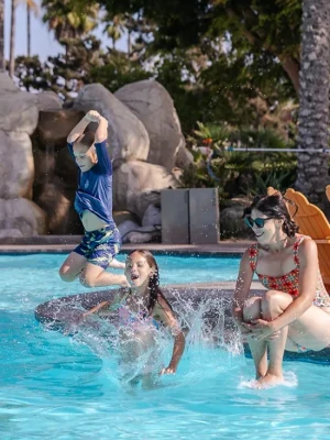 Three kids splash and jump into a sunny pool as a woman watches nearby, surrounded by rocks, palm trees, and sunny resort vibes.