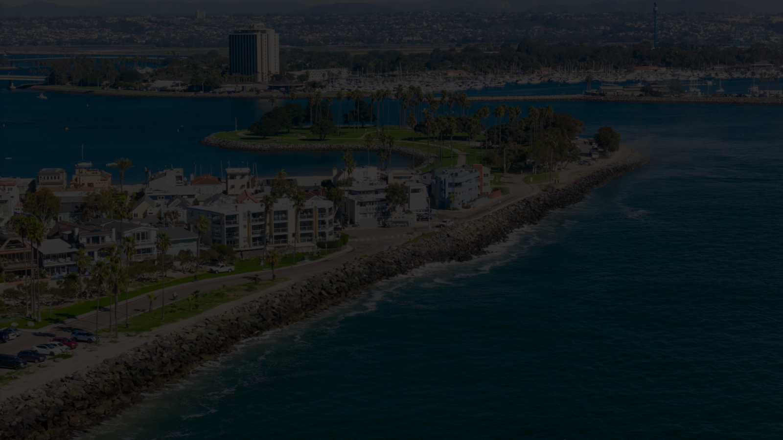 A coastal town with houses along a rocky shoreline, calm blue water, and distant buildings under a clear sky.