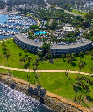 An aerial view of a curved, seaside hotel surrounded by a green lawn, palm trees, a winding path, and a marina with numerous boats nearby.