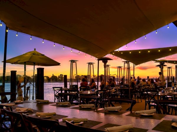 A lakeside restaurant at sunset with string lights, outdoor tables, umbrellas, and palm trees silhouettes against a colorful sky, diners eating.