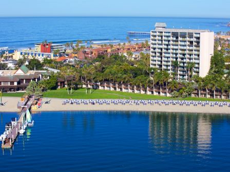 A sunny coastal resort with a sandy beach, palm trees, a dock, calm blue water, and a hotel building overlooking the sea, as people enjoy the shore.