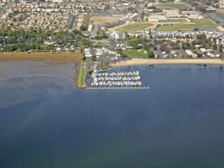An aerial view shows a coastal marina with numerous boats docked at a pier, adjacent to a neighborhood and grassy fields by the water.