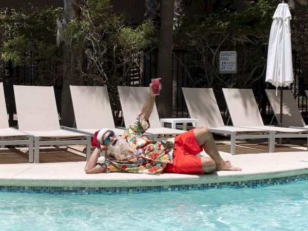 A person wearing colorful floral shirt and red shorts lounges by a pool, lying on their side with legs raised, sunbathing near pool chairs.