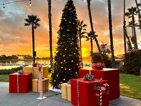 A decorated Christmas tree lit with colorful ornaments stands by wrapped gift boxes, palm trees, and warm sunset lights.