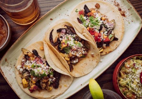 Three tacos on a platter with toppings like cheese, veggies, and herbs, plus a lime wedge and drinks nearby. End with a period.
