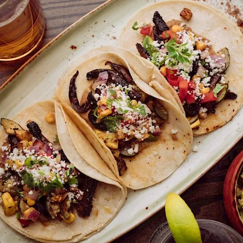 Three tacos on a platter with toppings like cheese, veggies, and herbs, plus a lime wedge and drinks nearby. End with a period.