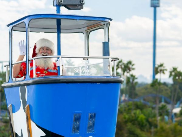 A blue Seaworld boat marked 22 carries Santa waving from the open cabin, outdoors with trees and sky in the background.