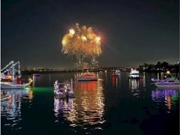 A nighttime river scene with colorful fireworks exploding above and reflections shimmering on the water beside lit boats.