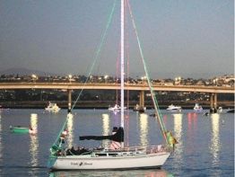 A sailboat with its mast raised is anchored on calm water near a shoreline at dusk, with lights glowing in the background.